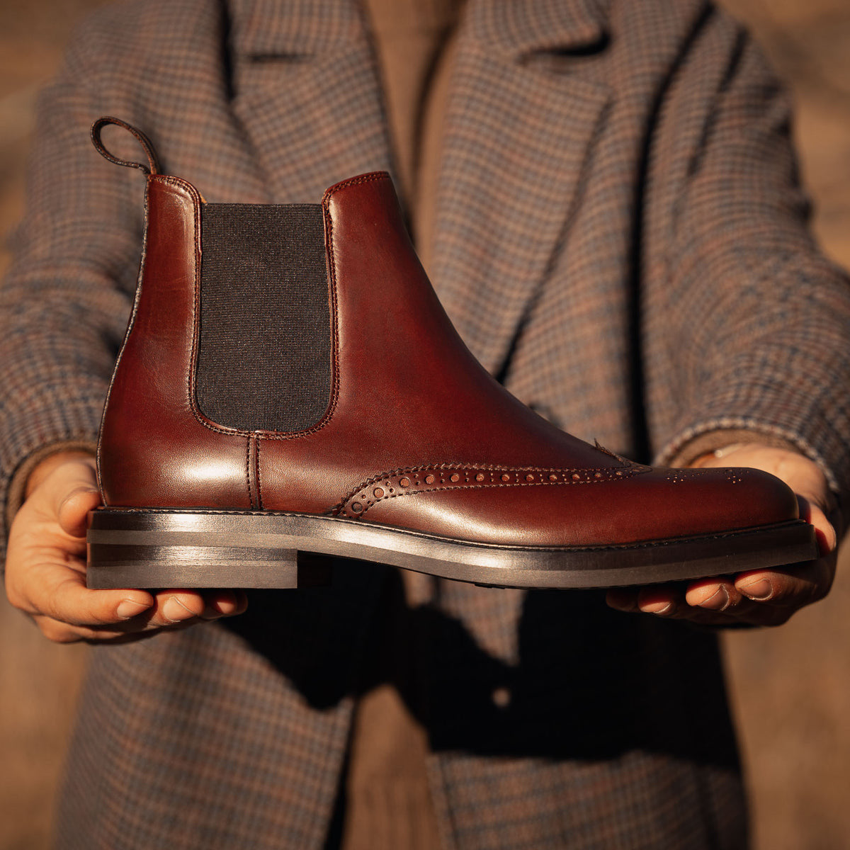 Brown Leather Brogue Chelsea Boot held in hands, showcasing fine craftsmanship, brogue detailing, and elastic side panels for a sleek and elegant design.